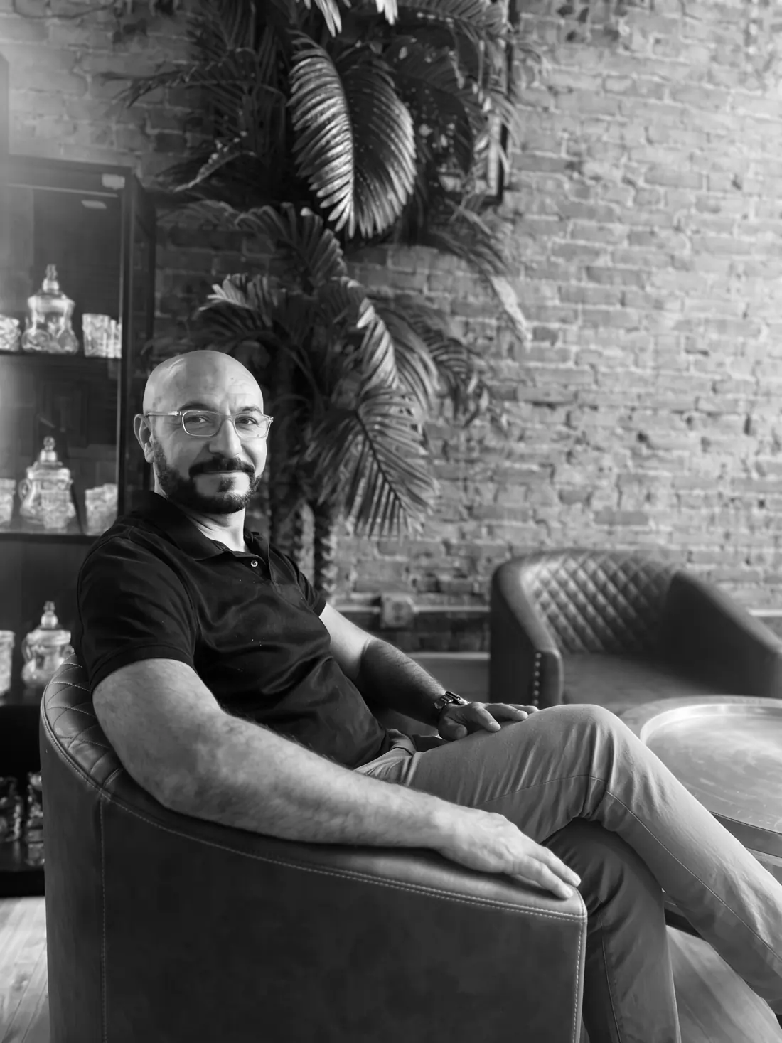 Joe Mikhail seated in an armchair in a coffee shop, black and white
